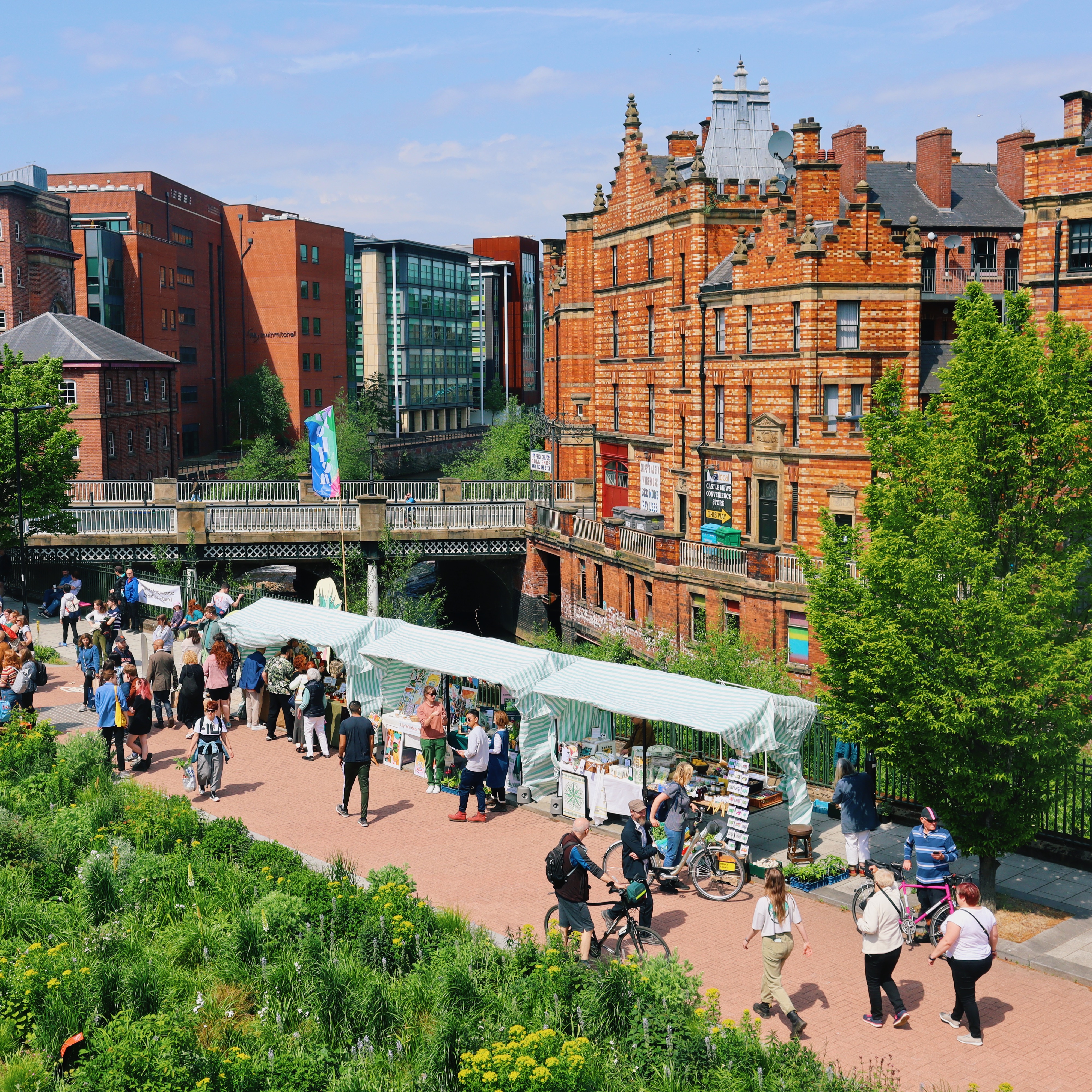 Outdoor market with white-striped stalls along a pedestrian walkway, surrounded by greenery. People are browsing and walking, some with bicycles. In the background, there is a large historic red-brick building with ornate architecture, alongside modern glass-fronted buildings.