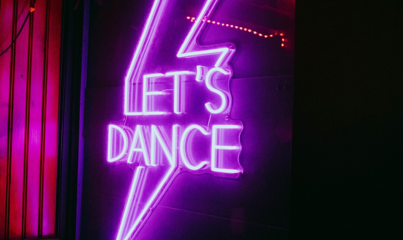 Pink and purple neon sign reading “Let’s Dance” inside a dimly lit bar or club space.