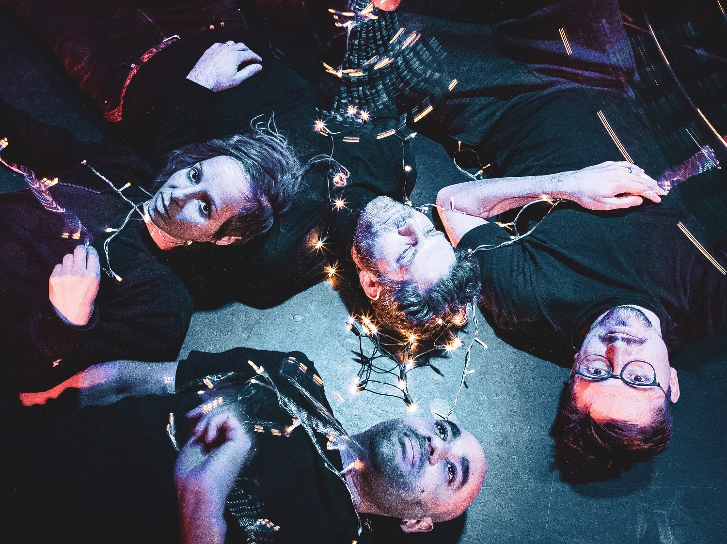 A dark an atmospheric photo of four people lying on the floor, staring upwards. There is a string of fairy light intertwined around them.