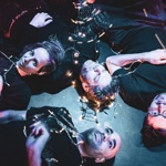 A dark an atmospheric photo of four people lying on the floor, staring upwards. There is a string of fairy light intertwined around them.