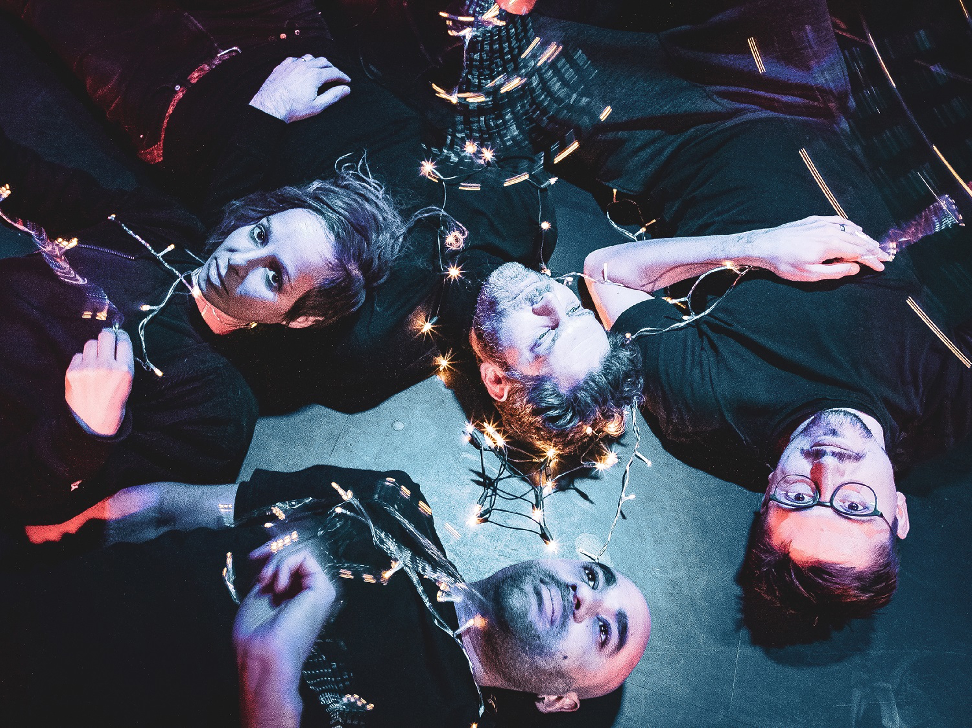 A dark an atmospheric photo of four people lying on the floor, staring upwards. There is a string of fairy light intertwined around them.