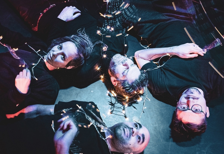 A dark an atmospheric photo of four people lying on the floor, staring upwards. There is a string of fairy light intertwined around them.