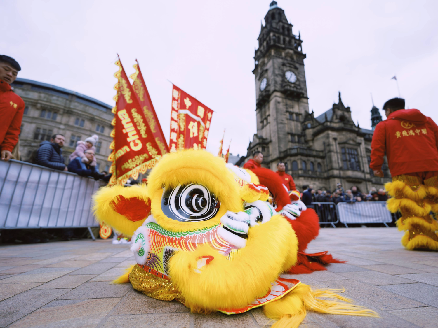 A close-up of a yellow lion dance costume lying on the ground, with intricate detailing and bright colors. Performers in red outfits and banners with Chinese characters are visible in the background, along with a historic building.
