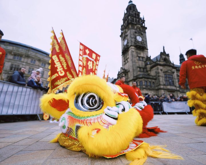 A close-up of a yellow lion dance costume lying on the ground, with intricate detailing and bright colors. Performers in red outfits and banners with Chinese characters are visible in the background, along with a historic building.
