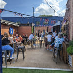 A lively outdoor courtyard on a sunny day with people gathered at high tables, bunting strung overhead, and brick buildings surrounding the space, creating a relaxed social atmosphere.
