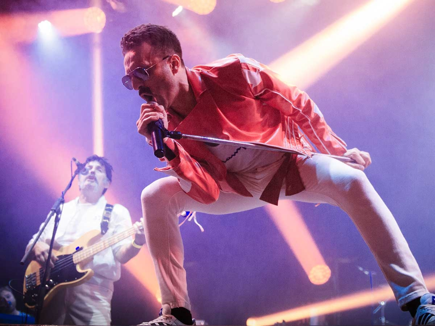 A dynamic concert scene with a performer in a bright red jacket and white pants leaning forward while gripping a microphone stand, illuminated by vibrant pink and orange stage lights. In the background, another musician plays an electric bass guitar, dressed in white, with additional lighting and equipment visible on stage.