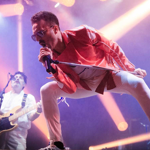 A dynamic concert scene with a performer in a bright red jacket and white pants leaning forward while gripping a microphone stand, illuminated by vibrant pink and orange stage lights. In the background, another musician plays an electric bass guitar, dressed in white, with additional lighting and equipment visible on stage.