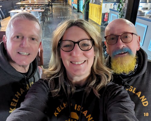 Three people wearing matching dark hoodies with yellow text reading ‘Kelham Island’ standing inside a spacious venue with wooden tables and chairs in the background.