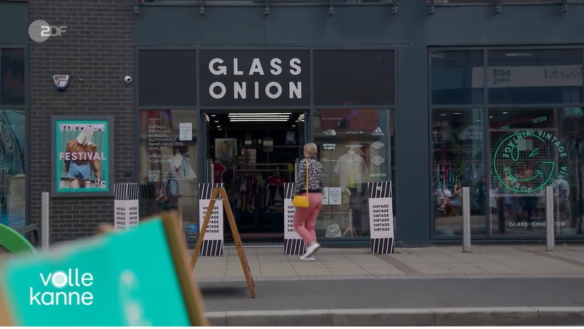 treet view of a clothing store with a black sign reading ‘GLASS ONION’ above the entrance. The shopfront has large glass windows displaying racks of clothes inside. A poster on the left advertises a festival, and the pavement outside is clear. Text on the bottom left reads ‘volle kanne.’