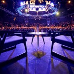 A wide auditorium filled with an audience seated in tiered rows, viewed from a stage setup that includes two upholstered chairs and a small table with two water bottles and glasses. Overhead rigging and circular lighting structures illuminate the venue, creating a dramatic atmosphere focused toward the stage area.