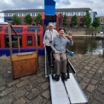 A person leaving the A&G Passenger Boat, in a wheelchair via a ramp. They are waving cheerfully at the camera.