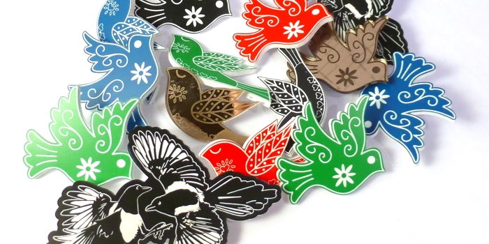 Collection of colorful bird-shaped brooches in red, green, blue, black, and metallic tones with intricate floral and leaf patterns, arranged on a white background.