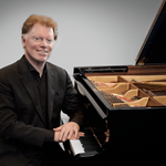 Classical pianist Tim Horton sits at a grand piano, smiling at the camera. He is dressed in a black suit with a black shirt.