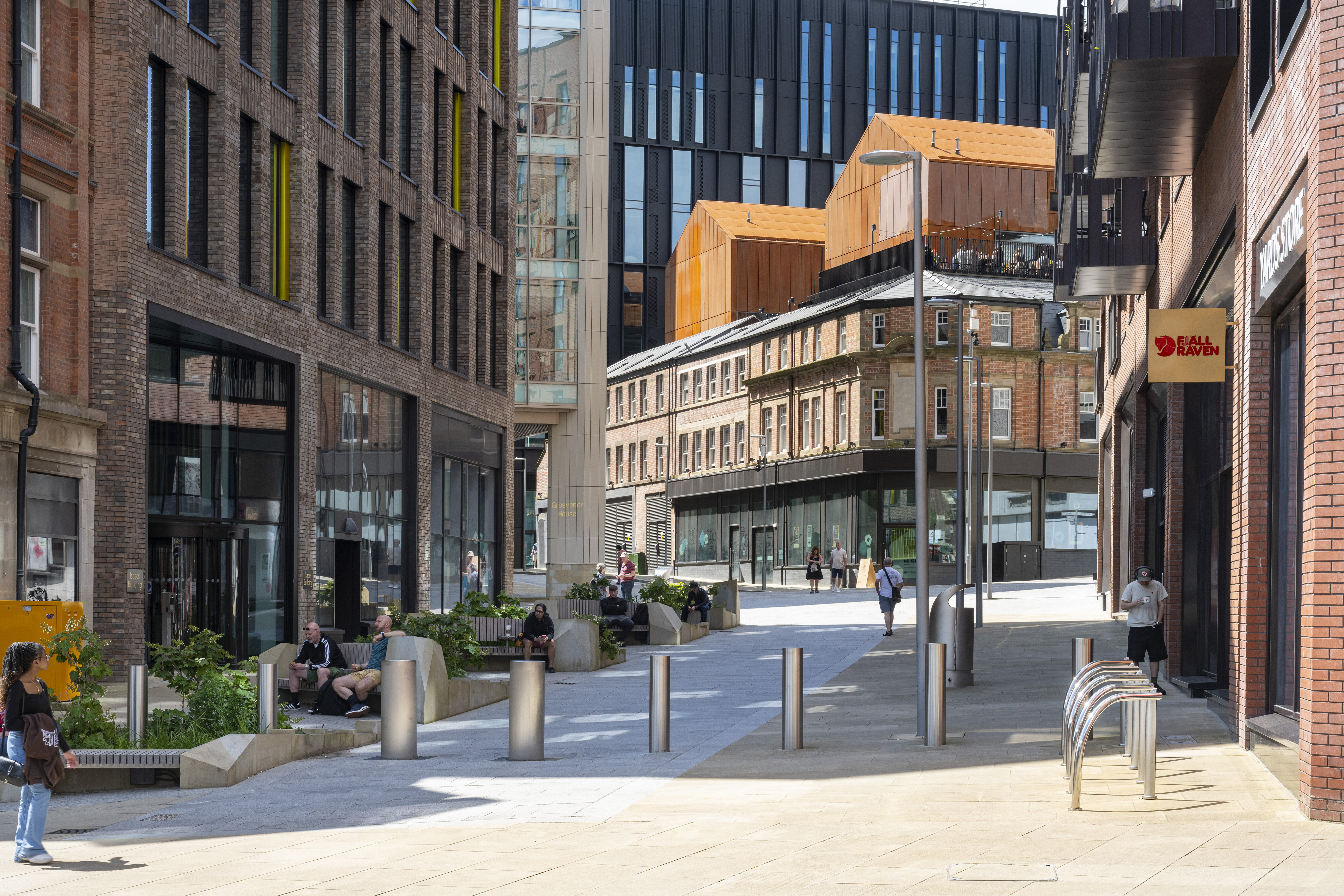 A pedestrianised shopping street in a city centre. People are strolling up and down, while others are sitting on benches.