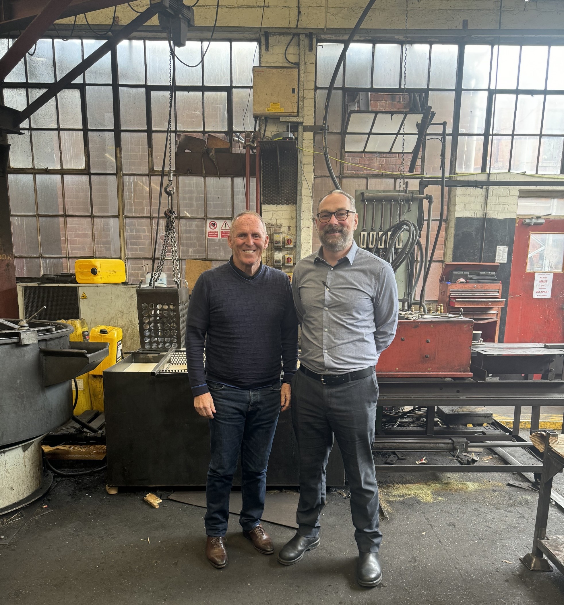 Two people standing inside an industrial workshop with large multi-pane windows in the background. The space contains heavy machinery, metal workstations, chains, and equipment, including a red tool cabinet and yellow safety blocks. The floor is dark and worn, and the overall setting suggests a manufacturing or engineering environment.