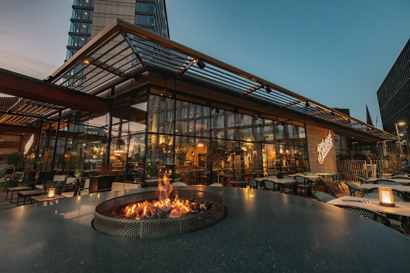 An evening view of a glass-walled restaurant surrounded by outdoor seating, with a circular fire pit lit in the foreground and soft lighting throughout the terrace.