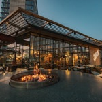 An evening view of a glass-walled restaurant surrounded by outdoor seating, with a circular fire pit lit in the foreground and soft lighting throughout the terrace.