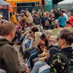 Rows of people sitting and eating food at a busy outdoor festival with stalls in the background.