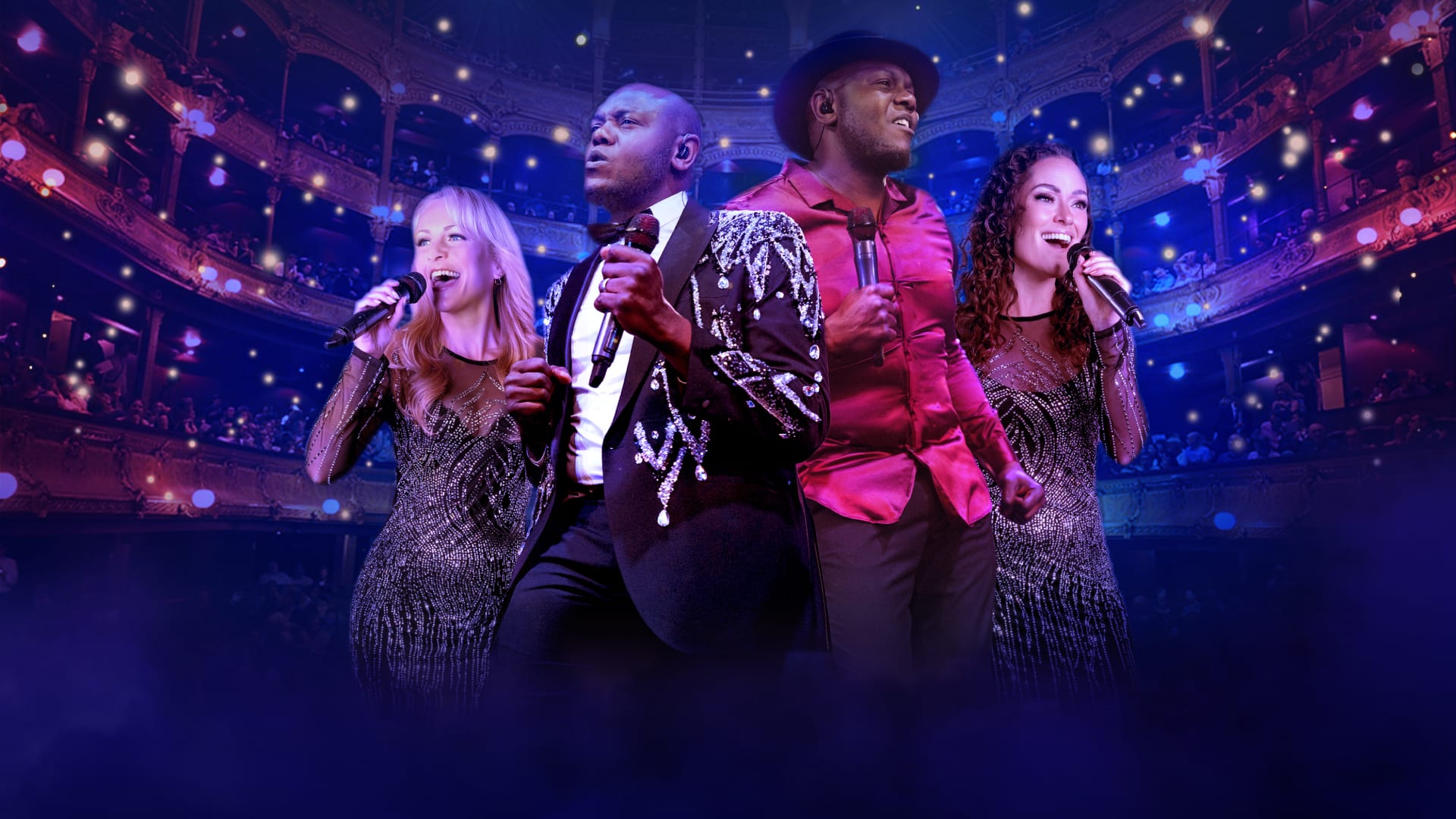 Four performers stand on stage holding microphones, dressed in sparkling and stylish outfits, including a sequined jacket and a bright red shirt. They are positioned in front of an ornate theatre interior with multiple balconies and glowing lights, creating a vibrant concert atmosphere with purple and blue stage lighting and mist in the foreground.