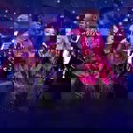 Four performers stand on stage holding microphones, dressed in sparkling and stylish outfits, including a sequined jacket and a bright red shirt. They are positioned in front of an ornate theatre interior with multiple balconies and glowing lights, creating a vibrant concert atmosphere with purple and blue stage lighting and mist in the foreground.