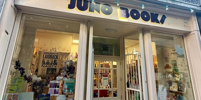 The exterior of an independent bookshop called “Juno Books,” featuring large front windows filled with posters and displays. The shopfront is cream-coloured with a central doorway leading inside, and a string of turquoise fairy lights hangs above the entrance.