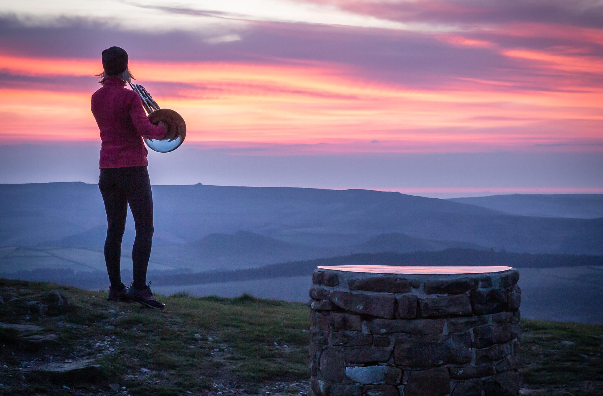 A woman stands on top of a hill in the Peak District, playing a French horn at sunrise.