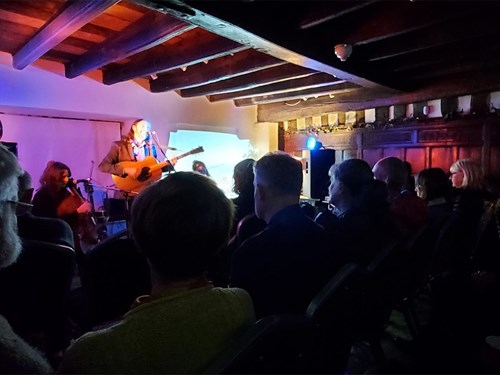 Indoor live music performance in a rustic venue with exposed wooden beams and warm lighting. A musician is playing an acoustic guitar and singing on a small stage, illuminated by blue and purple lights. A cello player is seated nearby. The audience is seated in rows of chairs, watching the performance. The background includes wooden panelling and a projector screen displaying a faint image.