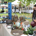 A pottery stall at an outdoor craft fair displays a variety of handmade ceramic items. A blue wooden shelf holds rows of bowls in different sizes and earthy tones. The table in front is covered with beige fabric and features large decorative vases, a patterned dish, small pots, and green plants. Leaflets and business cards are scattered on the table. In the background, white tents and other stalls are visible under leafy trees.