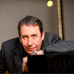 Jools Holland, dressed in a dark shirt and a grey wool jacket, leans on a black grand piano.