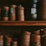 A wooden shelving unit is filled with neatly stacked handmade pottery bowls, cups, and lidded containers in earthy brown tones. The pieces vary in size and shape, with some marked by small handwritten labels indicating prices. The warm lighting highlights the smooth, natural texture of the ceramics arranged closely together on the shelves.