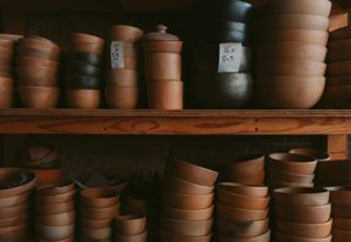 A wooden shelving unit is filled with neatly stacked handmade pottery bowls, cups, and lidded containers in earthy brown tones. The pieces vary in size and shape, with some marked by small handwritten labels indicating prices. The warm lighting highlights the smooth, natural texture of the ceramics arranged closely together on the shelves.