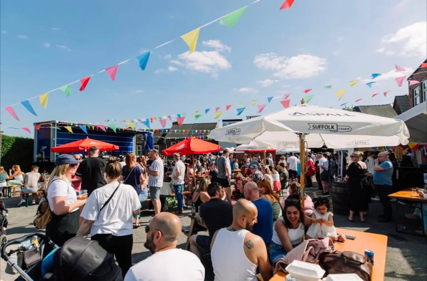  A lively outdoor event at a pub beer garden, with crowds sitting at picnic tables under colourful bunting and large umbrellas on a bright sunny day.
