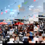  A lively outdoor event at a pub beer garden, with crowds sitting at picnic tables under colourful bunting and large umbrellas on a bright sunny day.