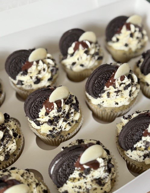 A box of vegan cupcakes decorated with swirls of cream frosting, crushed chocolate cookies, and whole Oreo biscuits. Each cupcake is topped with a drizzle of chocolate and a white chocolate button.