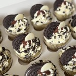 A box of vegan cupcakes decorated with swirls of cream frosting, crushed chocolate cookies, and whole Oreo biscuits. Each cupcake is topped with a drizzle of chocolate and a white chocolate button.