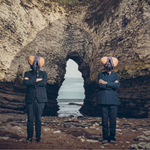 The two members of I Monster standing on a beech, wearing suits and fly head masks.