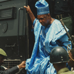 A person in African Traditional Dress is helped down from a large vehicle, surrounded by armed soldiers.