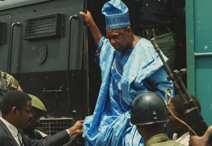 A person in African Traditional Dress is helped down from a large vehicle, surrounded by armed soldiers.