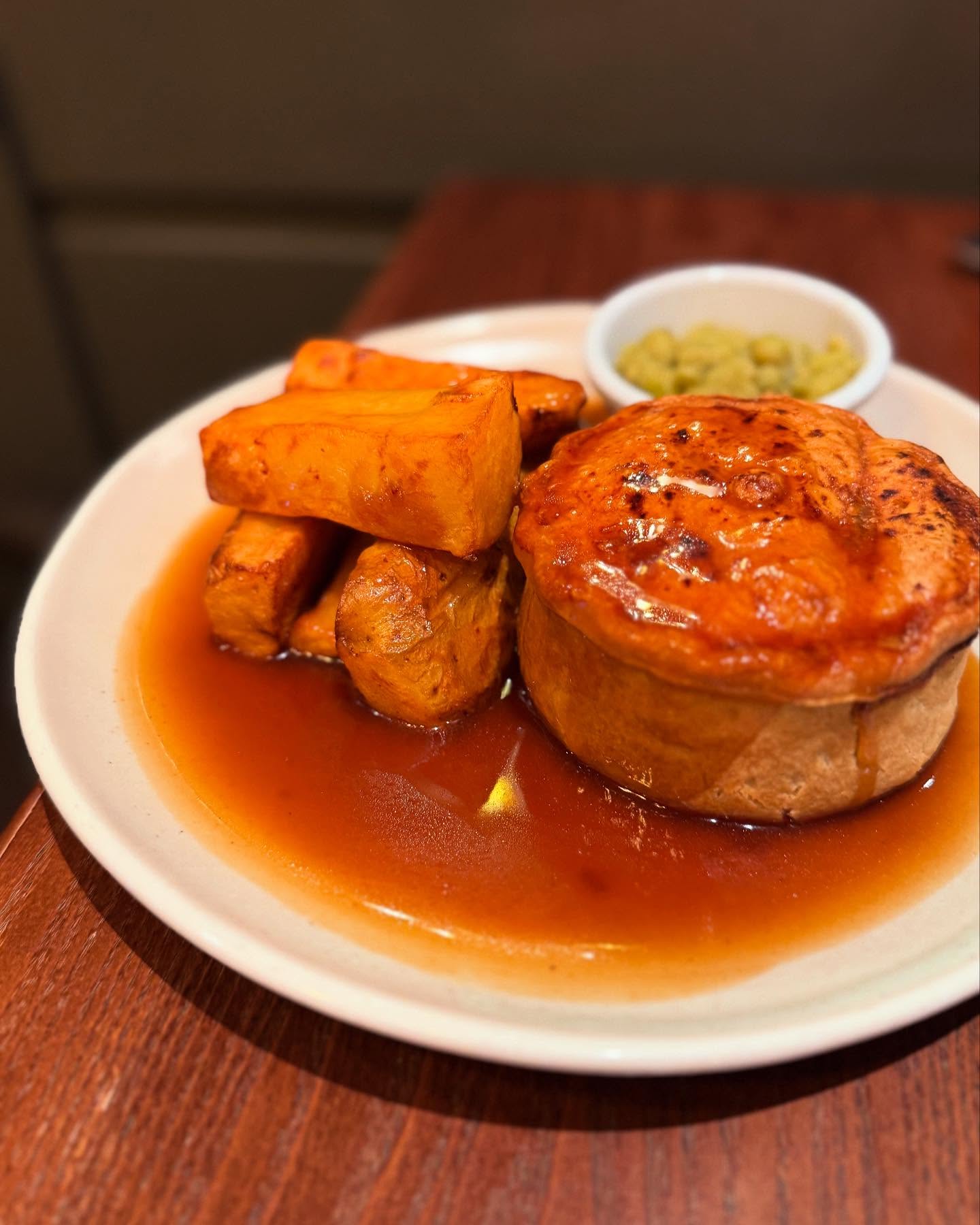A plated pub meal consisting of a golden pie served with thick-cut roasted potatoes, gravy, and a small bowl of mushy peas.
