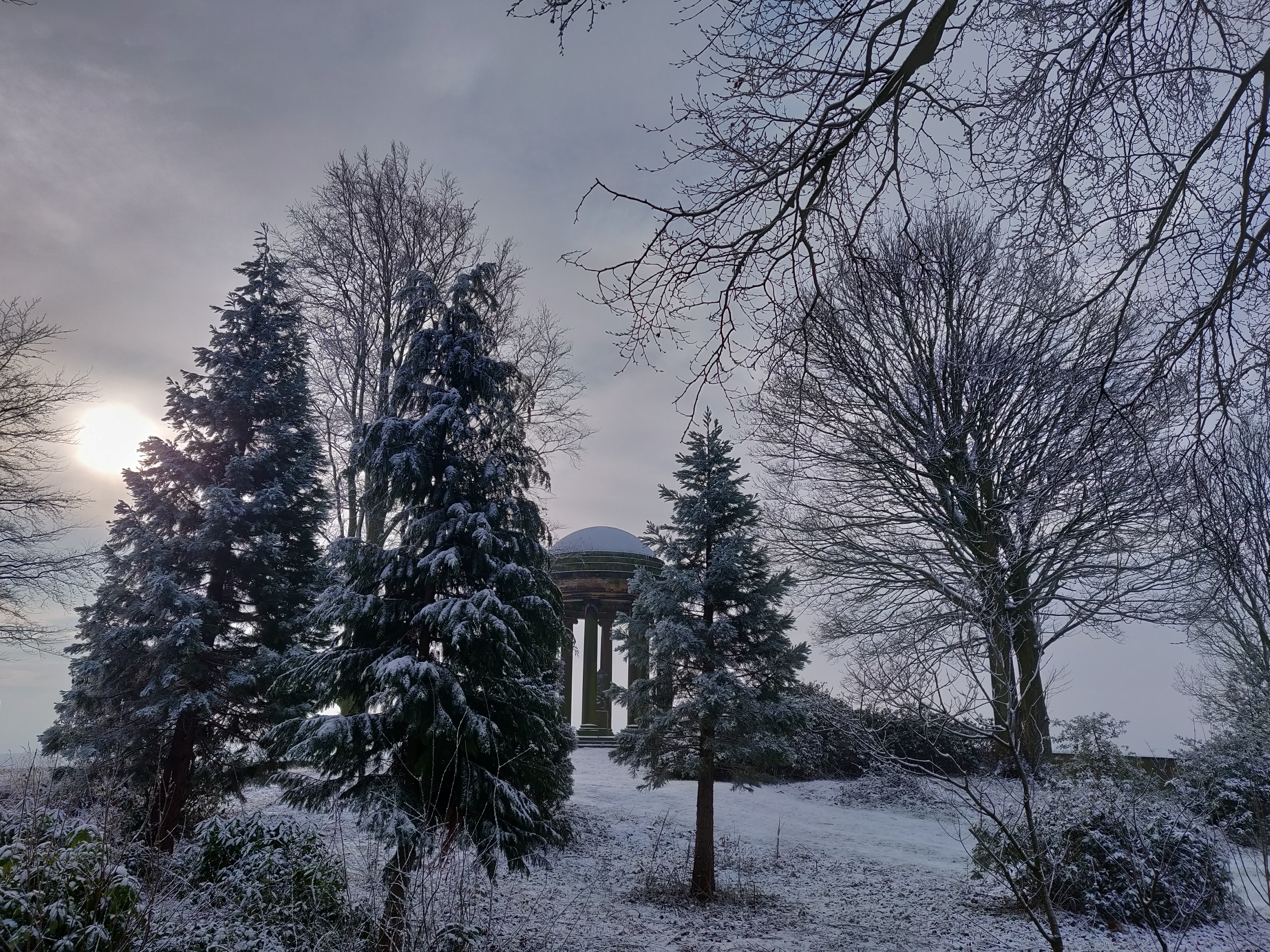 A winter landscape with snow-covered trees surrounding a small classical temple with columns. The low sun glows faintly through a cloudy sky, casting a soft light over the scene. Bare branches frame the view, and the ground and evergreens are dusted with fresh snow, creating a calm, wintry atmosphere.