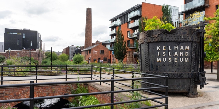 Outdoor view of Kelham Island Museum in Sheffield. A large industrial metal container with “Kelham Island Museum” written on it stands in the foreground near railings and a water channel. Surrounding the area are modern apartment buildings, a tall brick chimney, and greenery under a cloudy sky.