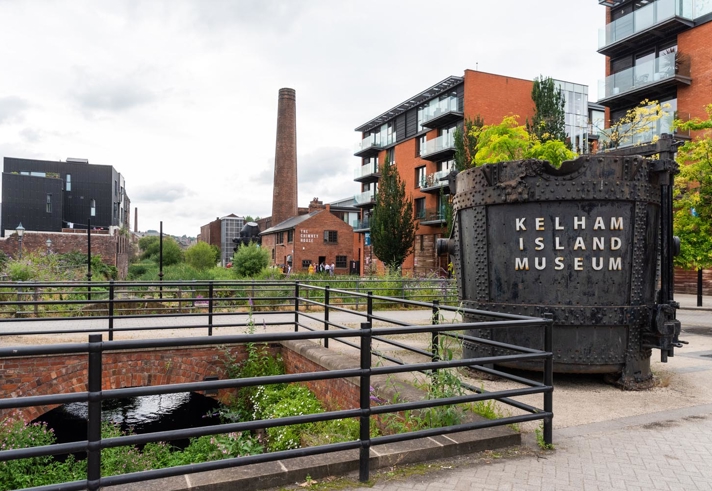 Outdoor view of Kelham Island Museum in Sheffield. A large industrial metal container with “Kelham Island Museum” written on it stands in the foreground near railings and a water channel. Surrounding the area are modern apartment buildings, a tall brick chimney, and greenery under a cloudy sky.