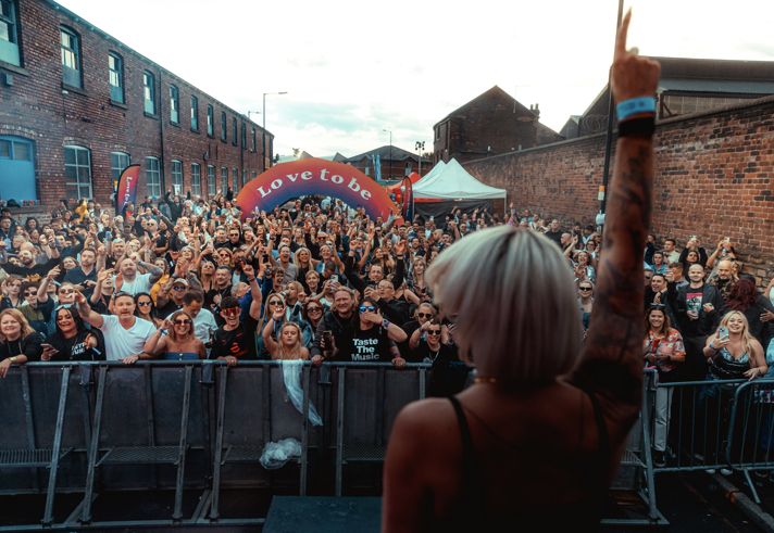 A performer on stage in front of a big crowd. In the middle of the audience is an arch with the words 'LOVE TO BE'.