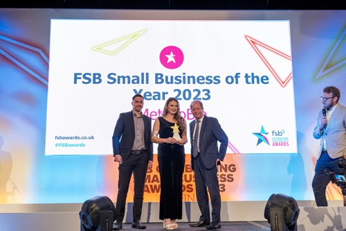 Three people standing on a stage at an awards ceremony with a large screen behind them displaying the text: “FSB Small Business of the Year 2023” and “fsbawards.co.uk #FSBAwards.” The person in the center is holding a trophy, while the two individuals on either side are dressed in formal attire. A fourth person with a microphone is positioned to the right, appearing to host or announce. The stage is brightly lit with colourful geometric designs in the background.