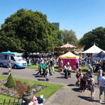 A busy outdoor festival scene in a park on a sunny day. Crowds of people walk and gather near colourful tents, including a bright yellow and pink stall, and a traditional carousel with a striped canopy. An ice cream van is parked on a paved path to the left. The foreground shows a flowerbed with neatly trimmed greenery, while large trees provide shade in the background.