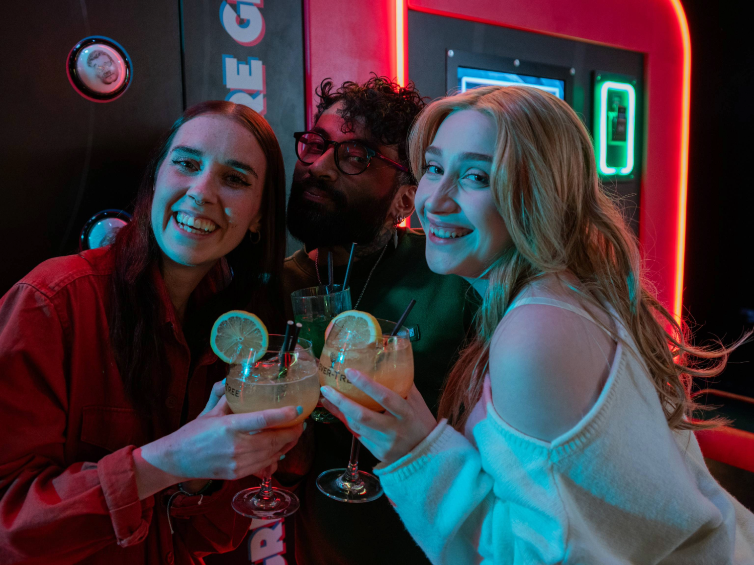 Three people standing close together in front of a neon-lit arcade game, each holding a cocktail glass with a light-coloured drink garnished with a slice of lemon and black straws. The background features bright red and green lights from the game machine, creating a vibrant and lively atmosphere. The group appears to be enjoying drinks while playing or watching the game.