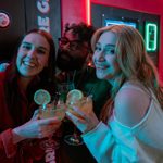Three people standing close together in front of a neon-lit arcade game, each holding a cocktail glass with a light-coloured drink garnished with a slice of lemon and black straws. The background features bright red and green lights from the game machine, creating a vibrant and lively atmosphere. The group appears to be enjoying drinks while playing or watching the game.