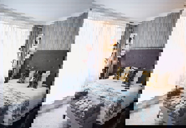 Elegant luxury bedroom featuring plush cushions on a king-sized bed, soft ambient lighting, and a large window with sheer curtains letting in natural light.