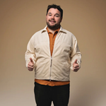 An almost full length photo of the performer, stood infront of a beige background. The performer is smiling and doing the thumbs up sign with both of his hands. 