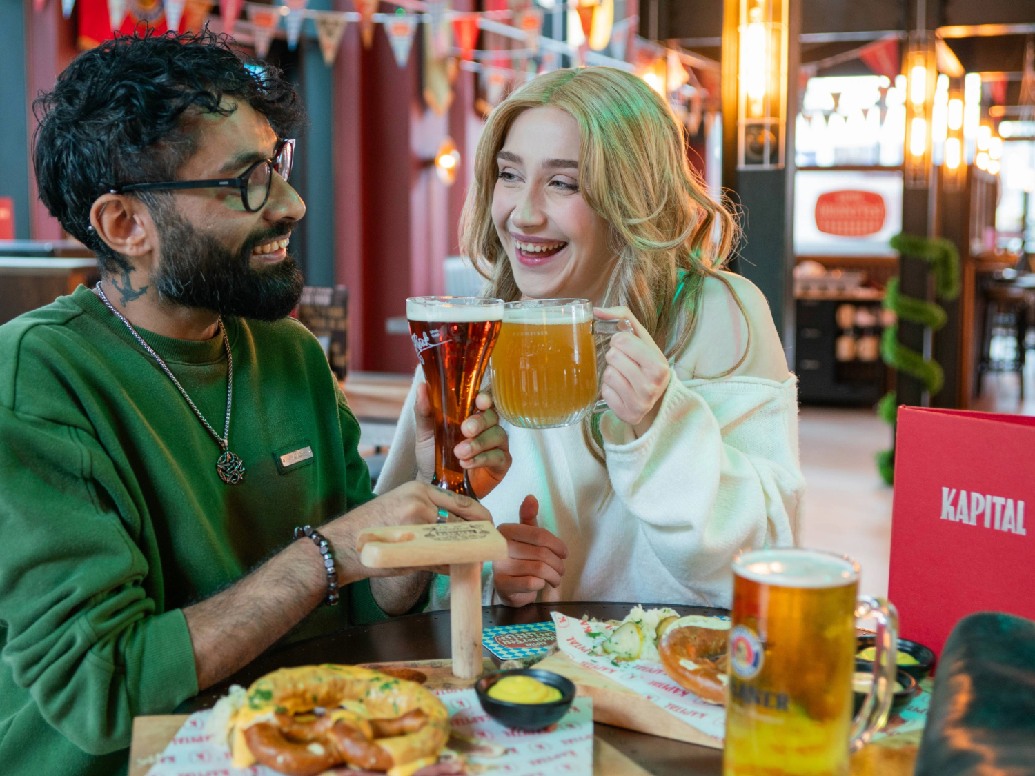 Two people are seated at a wooden table in a lively bar or beer hall, holding large glasses of beer and ale in a toast. The table has plates of food, including pretzels with mustard dip and other snacks. A red menu with the word “KAPITAL” is visible, and the background features colourful bunting, warm lighting, and a modern interior design.